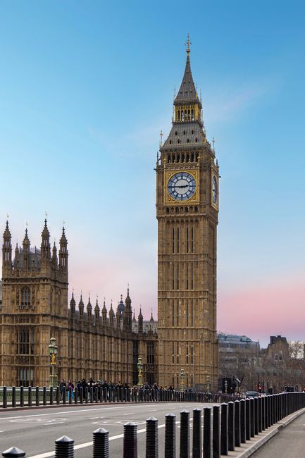 The image depicts London's iconic Big Ben clock tower and the Palace of Westminster fronted by a paved roadway with black metal bollards lining the edge. The tower's ornate Gothic architecture is prominent, with detailed stonework and spires, topped by a spire with a clock face showing the time as approximately 4:25. The sky above is clear with a soft gradient of blue and pink hues, indicating either early morning or late afternoon. To the left, part of the palace's facade with elaborate stone carvings and turrets is visible, all appearing clean and well-maintained. The scene is bathed in natural light, highlighting the intricate architectural details and the historical significance of the landmark. This setting emphasizes the importance of surface cleaning and maintenance to preserve the appearance of historic structures, much like how Carpet Cleaning Westminster delivers surface cleaning and sanitisation services for homes and businesses in Westminster, SW1.