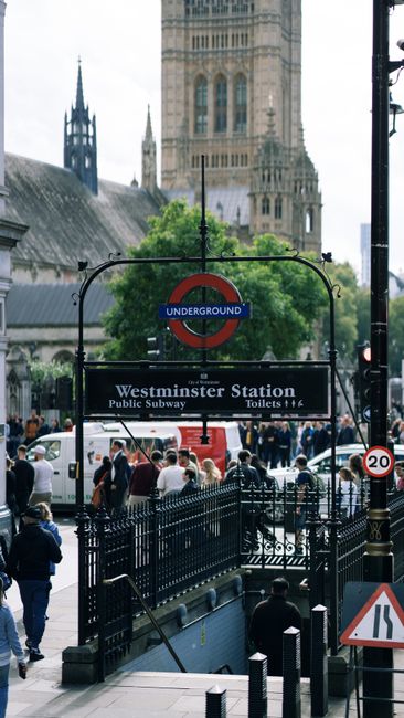 An exterior view of Westminster Station entrance captured during daytime with natural lighting. In the foreground, there is a black metal railing surrounding the station steps leading underground. The station sign displays the iconic roundel logo with the name 'Westminster Station' and additional information about public subway and toilets. The background features London’s historic architecture, including the gothic-style Westminster Abbey with tall spires and intricate stonework, set against a cloudy sky. Pedestrians, dressed in casual clothing, are seen walking on the busy sidewalk, with some arranging themselves near the entrance. The scene emphasizes the urban environment typical of Westminster, focusing on public accessibility and transportation hub surroundings without any surface cleaning or sanitisation elements visible.