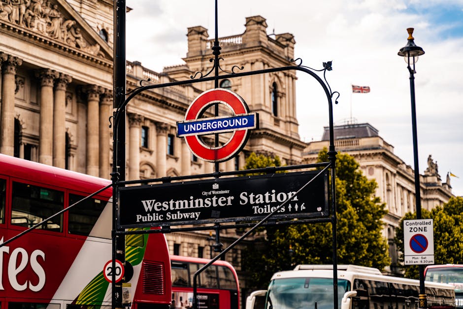 Close-up of a black and red underground sign for Westminster Station, situated in front of historic classical architecture with ornate columns and detailed stonework. The sign indicates it is a public subway station in Westminster, London, with a partly cloudy sky and trees visible in the background. Nearby, a red double-decker bus and some white vehicles are seen parked or moving. Street lamps and a controlled zone sign are also visible, contributing to the urban setting. The area is well-lit with natural daylight, highlighting the detailed facades of the buildings and the signage details, providing a clear view of the station entrance and surrounding cityscape.